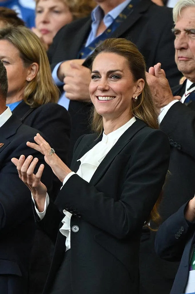 Person clapping, wearing a stylish suit with a ruffled blouse, amidst a crowd at an event