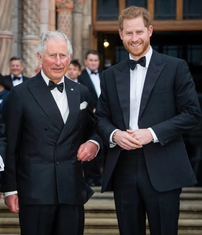 King Charles and Prince Harry in tuxedos stand arm-in-arm at an outdoor event, smiling for the camera
