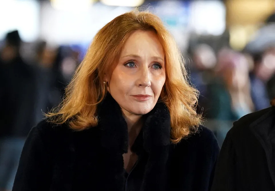 A person with long hair is wearing a dark coat, standing in a busy, blurred background setting