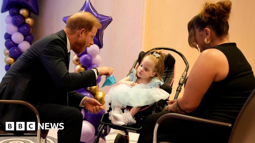 Prince Harry presents Stoke-on-Trent girl with WellChild Award Prince Harry in a dark suit sits besides a little girl wearing a light blue dress. A woman in a black top sits next to the pair.