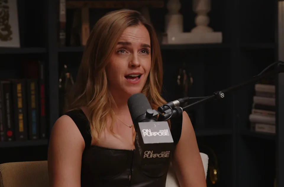 Person speaking into a microphone during a podcast. They are wearing a sleeveless outfit. Shelves with books and decor are visible in the background