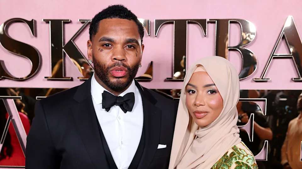 A man in a tuxedo and a woman in a beige hijab pose together at an event with a pink backdrop.