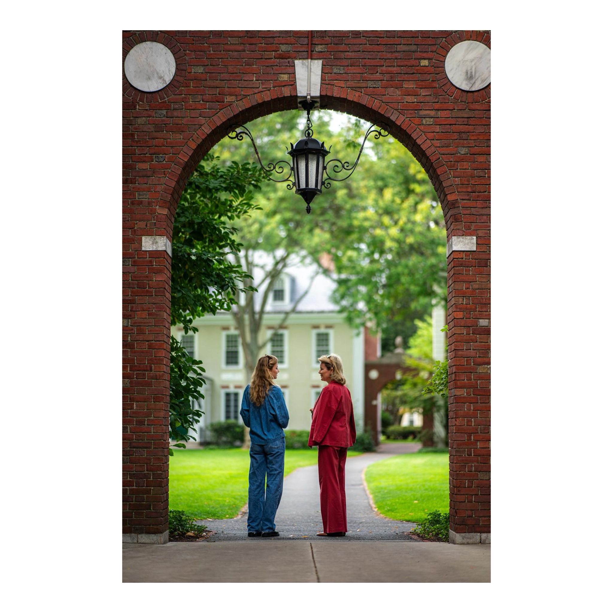 Princess Elisabeth wearing jeans and Queen Mathilde in a pink suit posing under an archway at Harvard