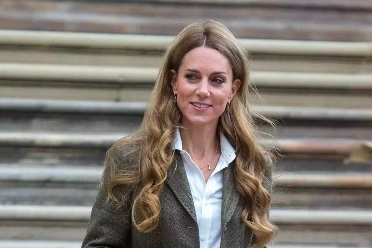 Long, wavy-haired woman in a brown jacket and white jersey standing in front of some stairs