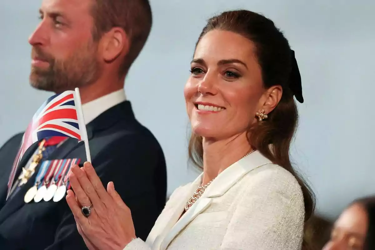A smiling woman holds a small United Kingdom flag while standing next to a man with medals on his suit.