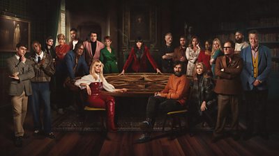 The Celebrity Traitors contestants sit and stand around a round table, which host Claudia Winkleman leans on.