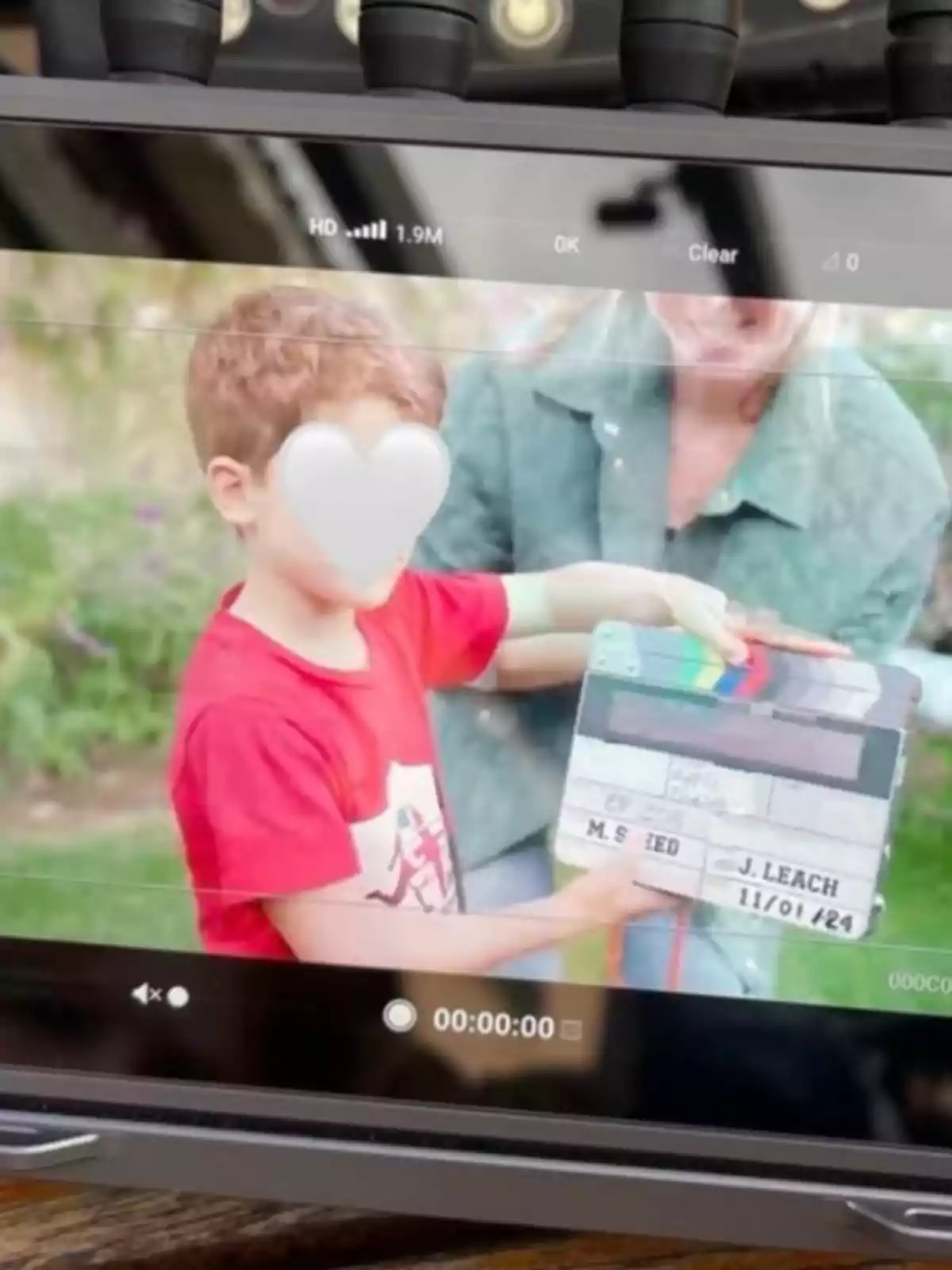 A boy in a red jersey holds a film clapperboard while an adult accompanies him outdoors; both appear on a recording monitor screen.