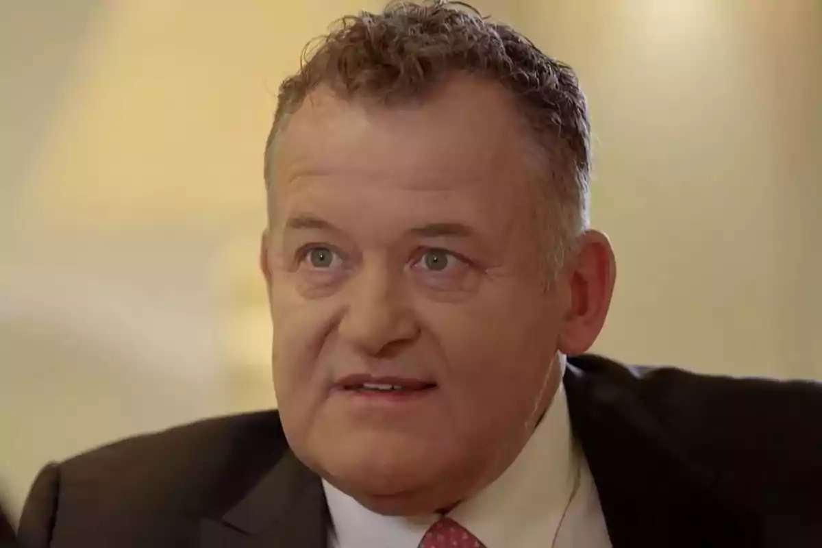 Paul Burrell in a dark suit and pink tie looking straight ahead with a serious expression. Screenshot from 'Viajando con Chester'