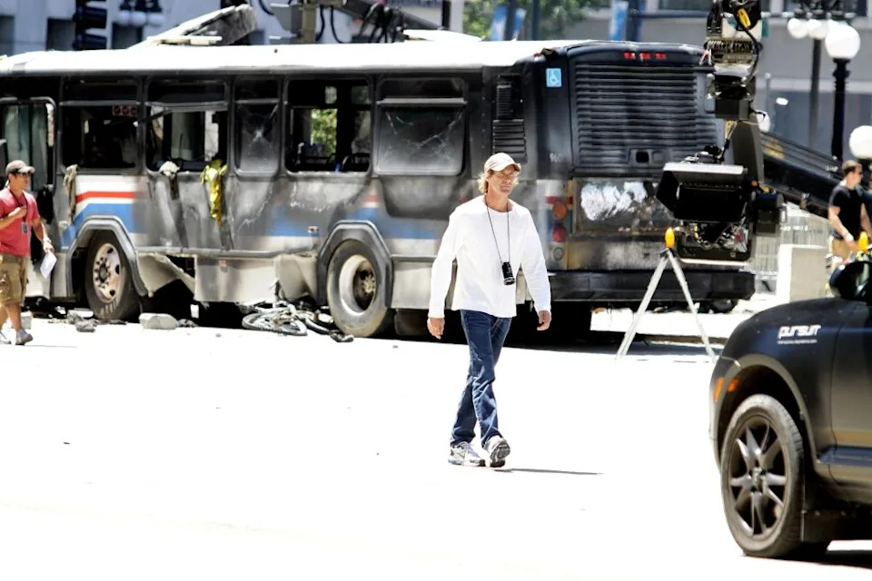 A person in casual clothing walks past a damaged bus on a film set, with crew members and equipment visible in the background