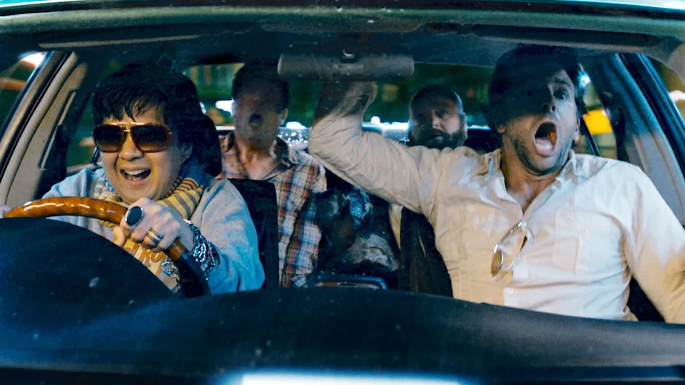 Four people in a car, reacting with shock and excitement. The driver wears sunglasses and a scarf