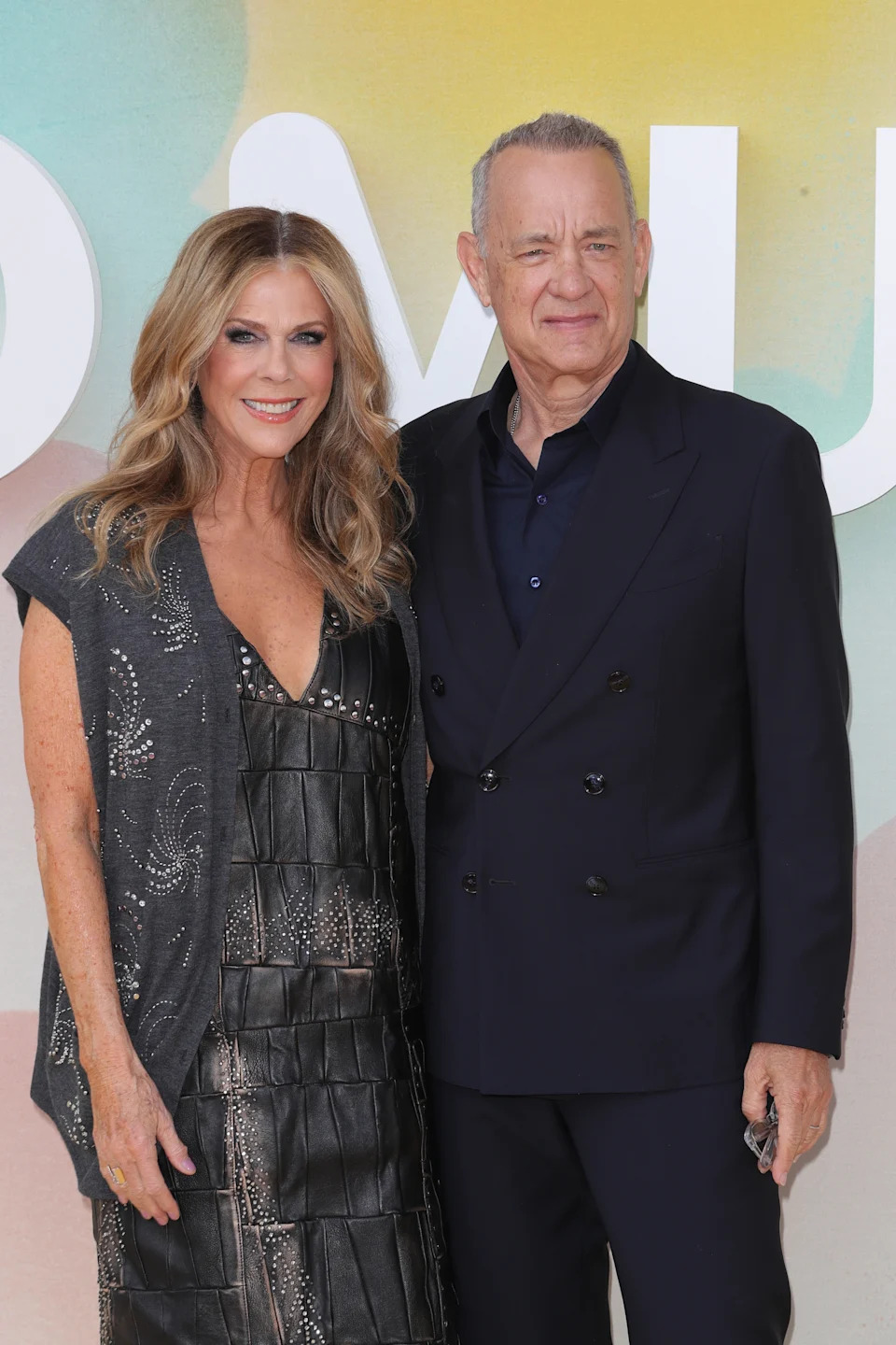 Two individuals smiling at a public event; one wears a sleeveless, textured dress, and the other is in a dark suit