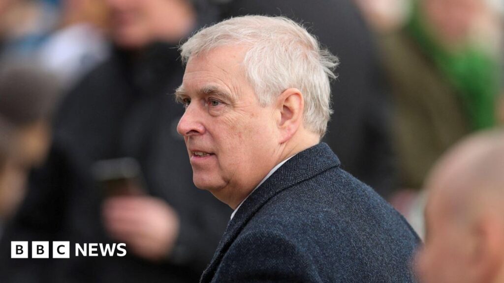 Prince Andrew wearing a dark overcoat over a white shirt, standing outdoors among a group of people. The background shows blurred figures