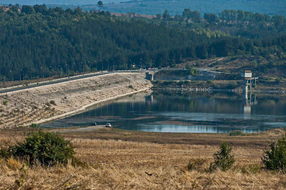 A serene landscape with a dam, calm water reflecting trees and sky, surrounded by hills and greenery