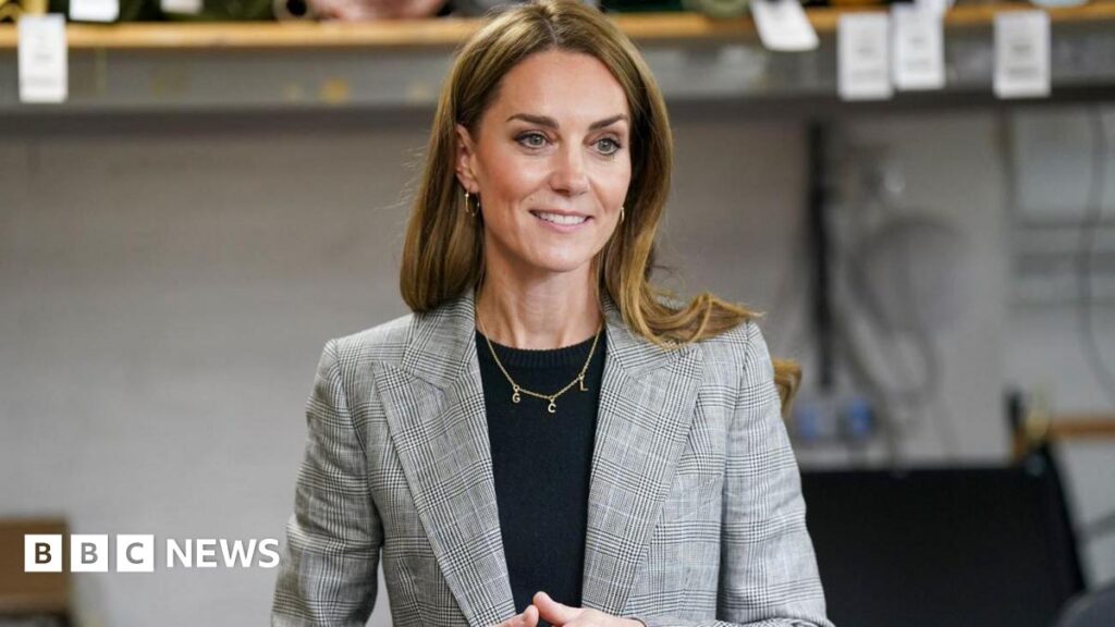 Catherine, Princess of Wales with long brown hair is wearing a grey and black blazer and black top and a gold necklace with the letters 'G, C, L' on it with small gold hoop earrings. She is smiling to the side with her hands clasped together. The background is blurred.