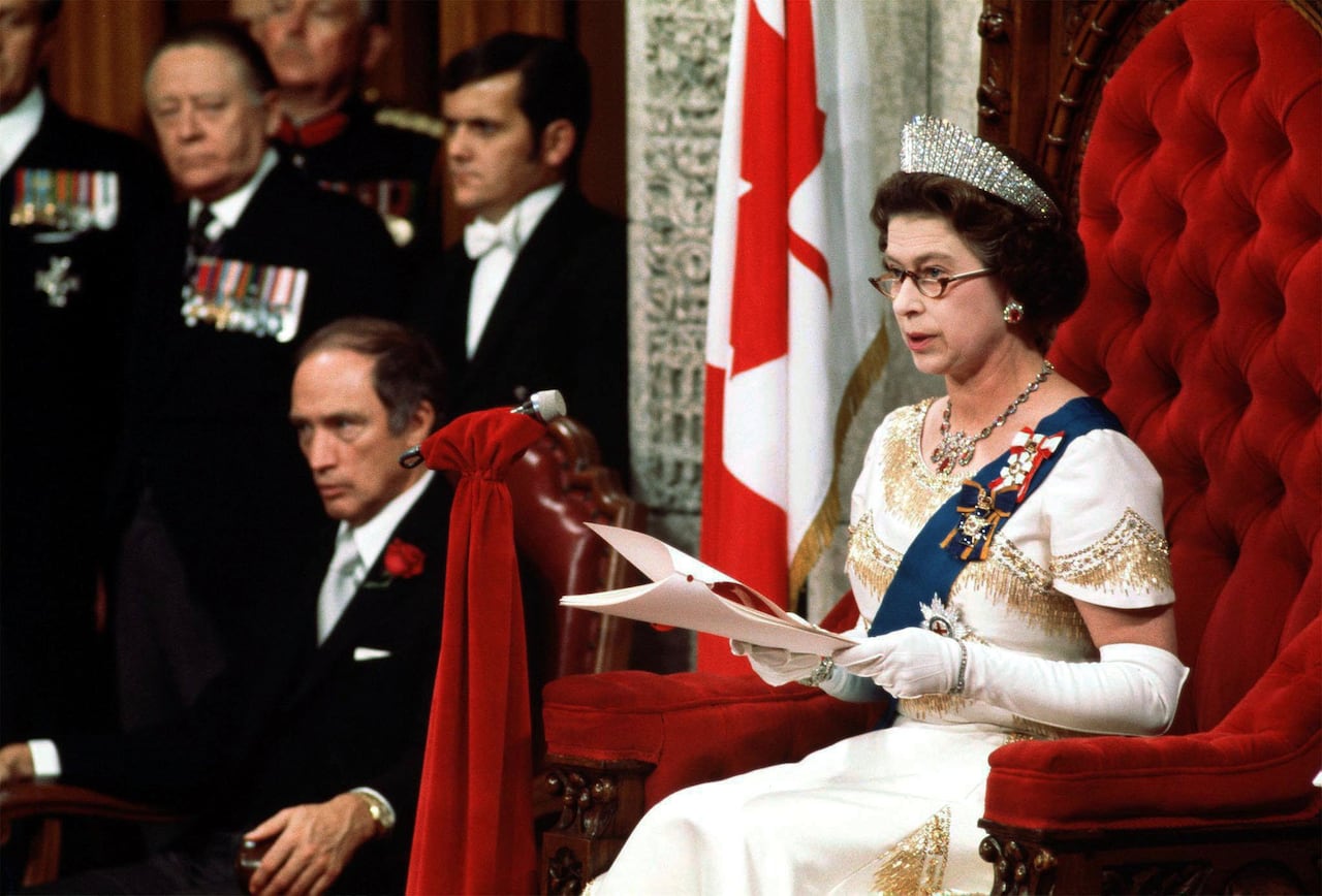 A woman wearing a crown, and sitting on an elaborate chair, reads a speech while other people look on.