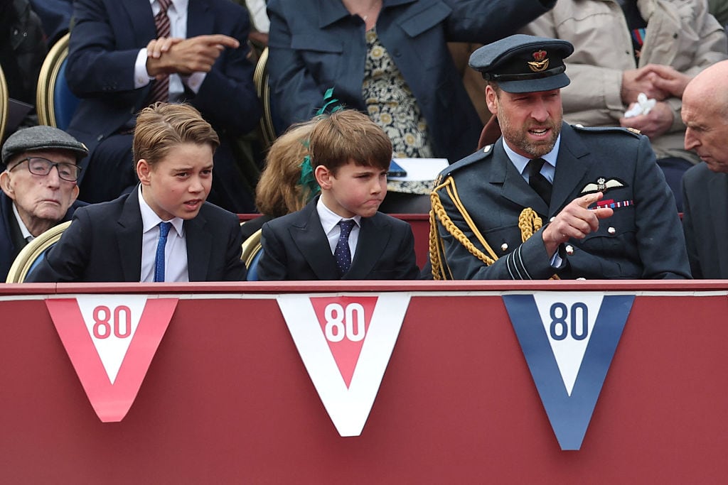 Two children wearing suits sit beside an adult in a military uniform behind a red wall that has decoractive Vs with the numeral 80 in them strung along its top.