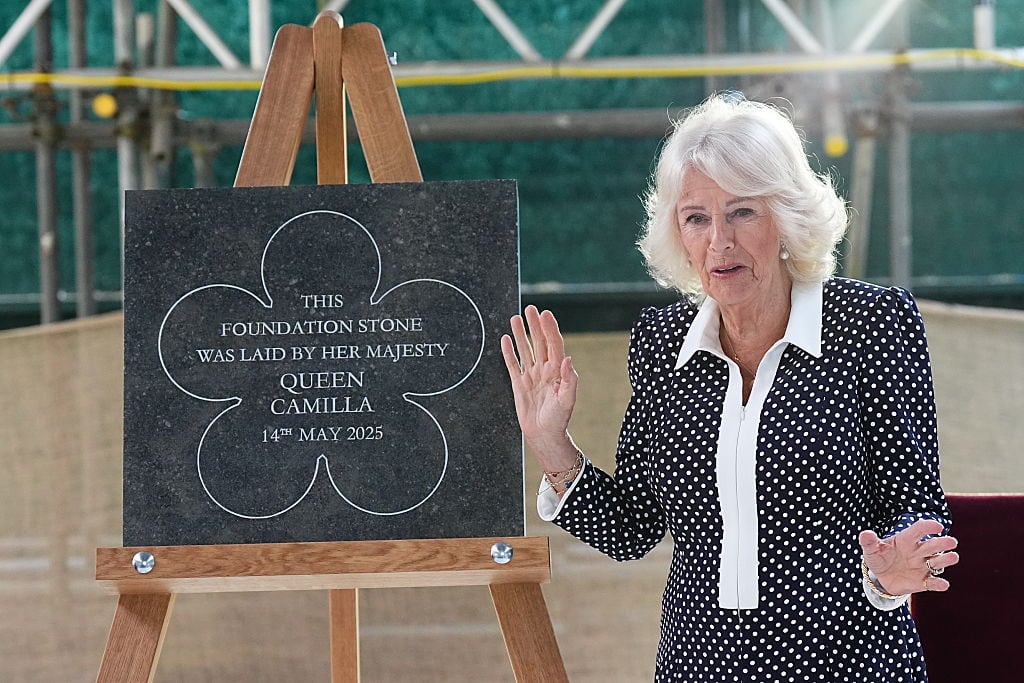 A person stands beside a square piece of stone displayed on an easel that has a floral-like etching around the words: This foundation stone was laid by Her Majesty Queen Camilla 14th May 2025.