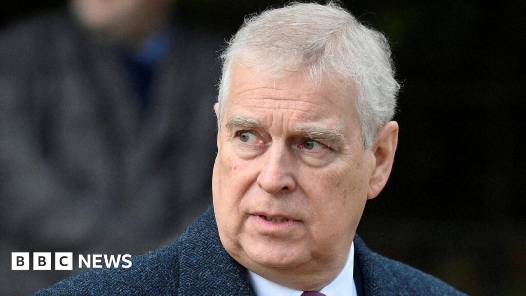 Prince Andrew wearing a dark textured overcoat over a white shirt and red tie, standing outdoors with a blurred background