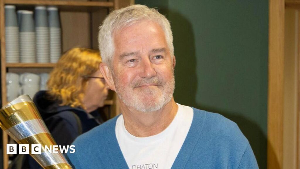 A man with grey hair and a grey beard holding a silver and gold baton while smiling. He is wearing a blue cardigan over a white round-kneck T-shirt.