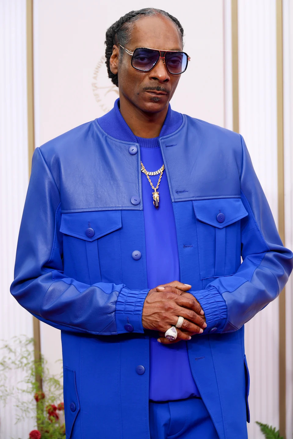 Person in a stylish blue jacket and shirt, wearing sunglasses and a gold necklace, poses confidently