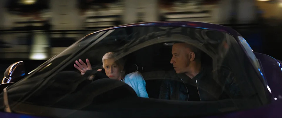Two people in a moving car, one gesturing animatedly, the other focused on driving; the scene suggests a dynamic conversation
