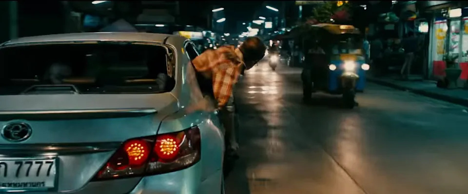 Person leans out of a moving car's window on a busy, evening street with vehicles and shops in the background