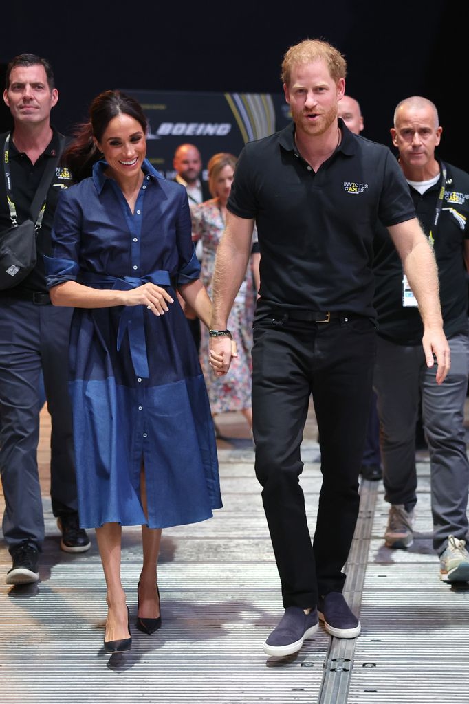 Meghan Markle and Prince Harry, Duke of Sussex walk hand in hand. Meghan is wearing a blue chambray shirt dress tied at the waist and Harry is wearing a black polo shirt and trousers. 