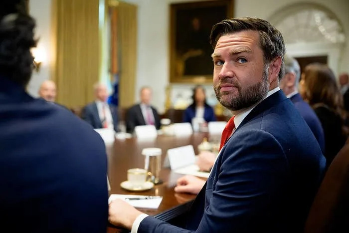 A person in a suit sits at a conference table in a formal meeting room, appearing attentive with others blurred in the background