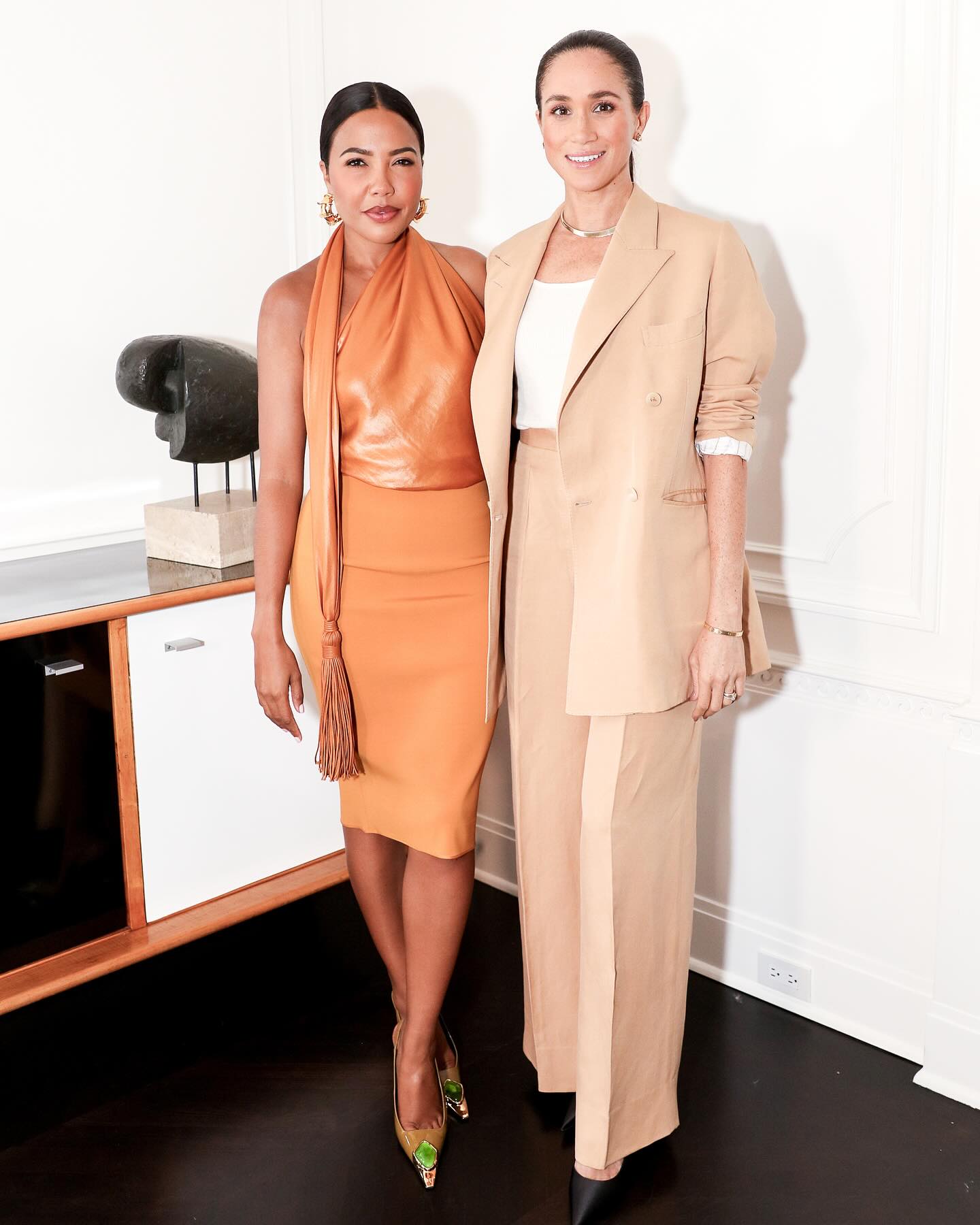 Meghan Markle wearing a beige suit standing next to Emma Grede, in an orange dress