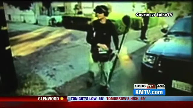 A person holding equipment walks past a car on a street. TV broadcast graphics show local weather and KMTV channel information