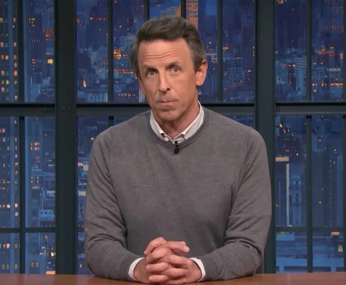 A person on a TV show set sits at a desk, wearing a gray sweater over a collared shirt, with a cityscape visible through the window behind them