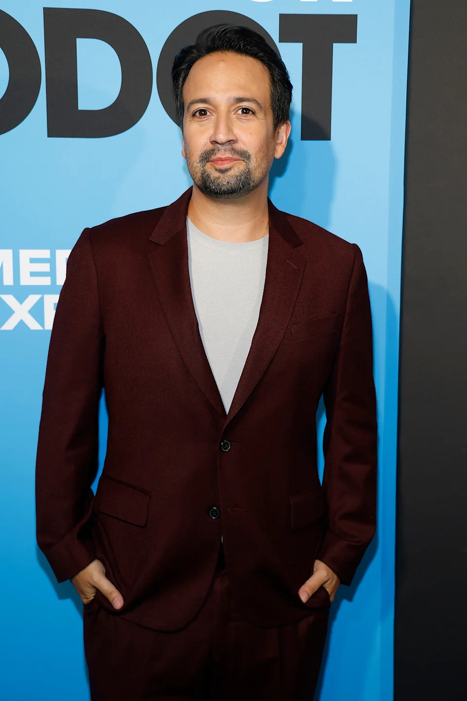 Person in a tailored dark suit and light shirt stands confidently, hands in pockets, at a media event backdrop