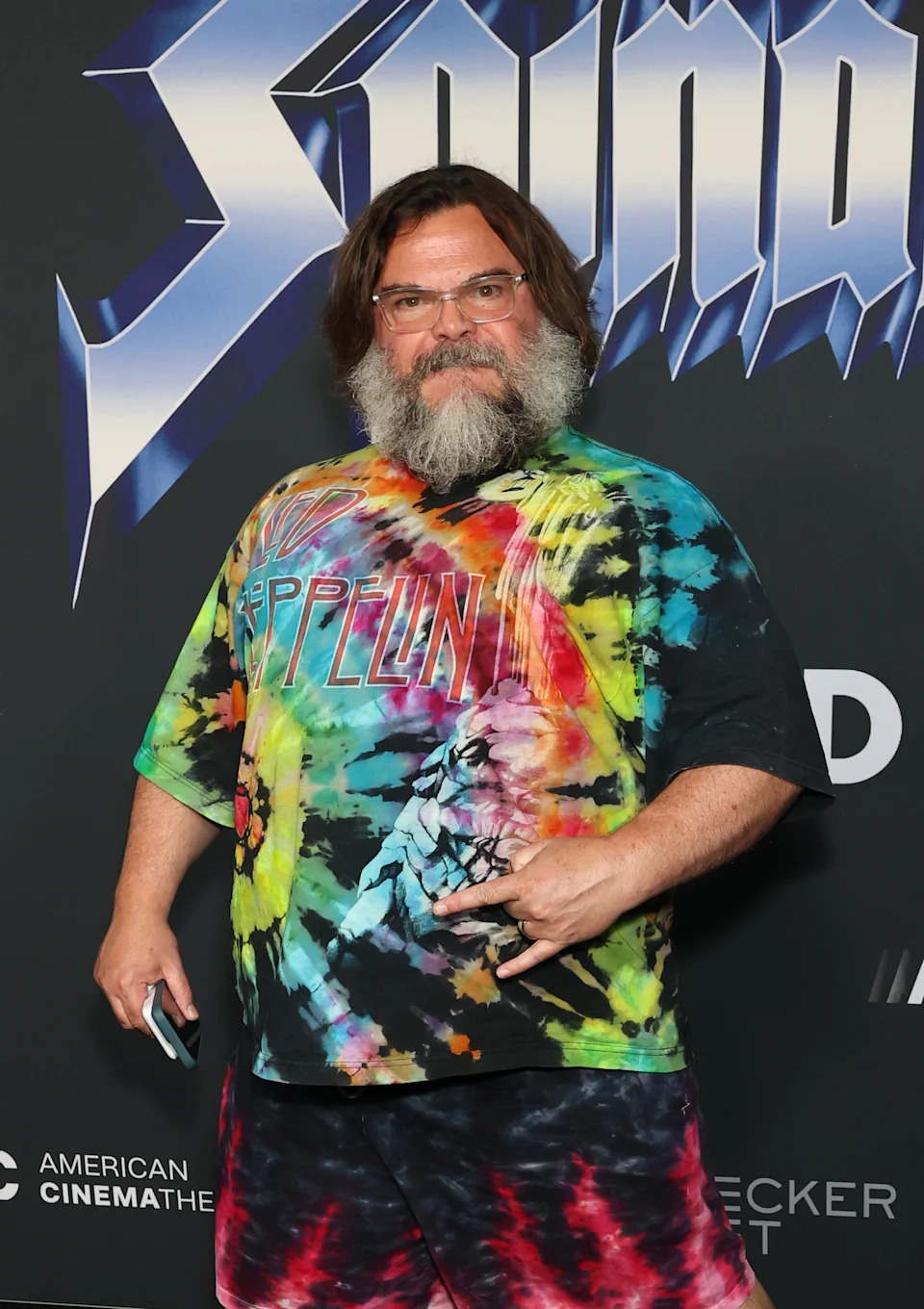 Celebrity posing on the red carpet, wearing a colorful tie-dye Led Zeppelin shirt and matching shorts, making a rock hand gesture