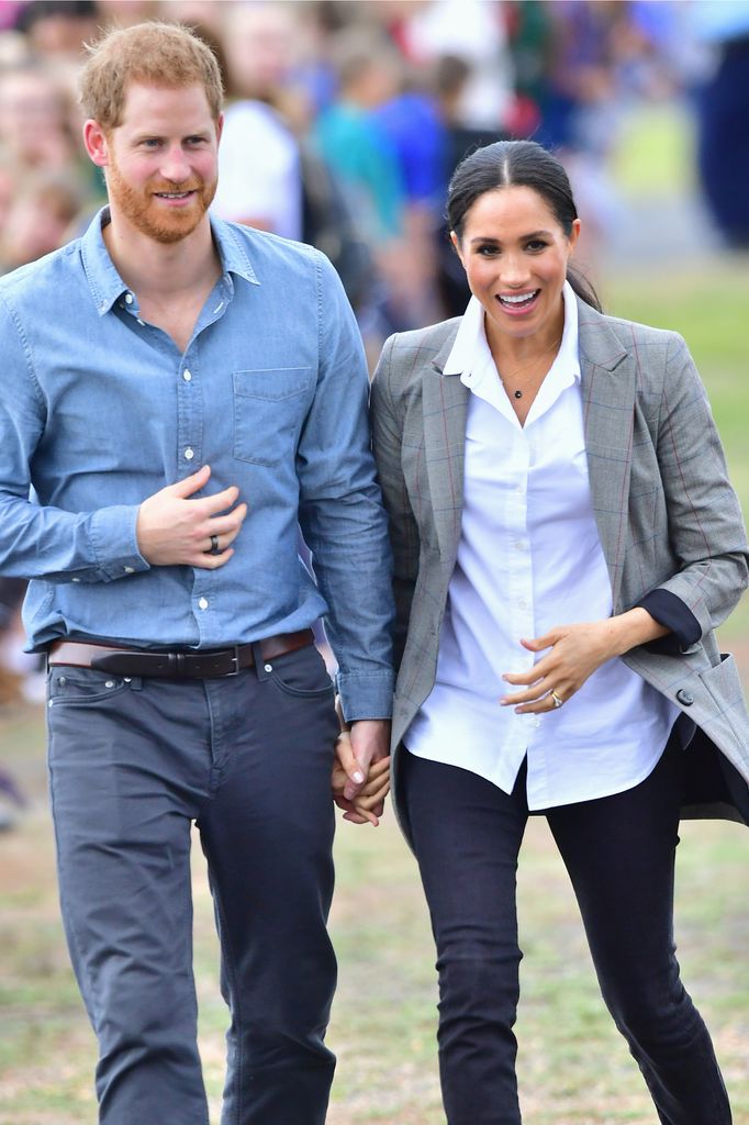 Prince Harry and Meghan Markle walk hand in hand. She is wearing a white shirt, black jeans and a grey blazer, he is wearing a blue shirt and chinos. 