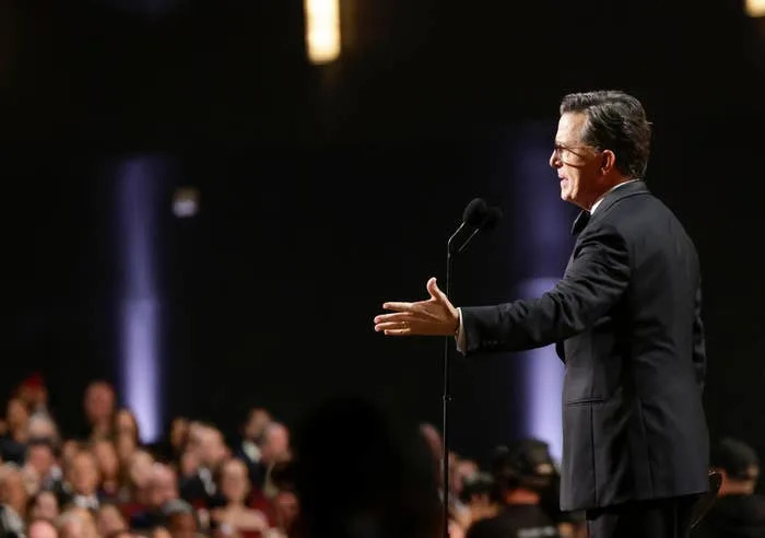 Stephen Colbert in a suit speaks into a microphone on stage before an audience at an event