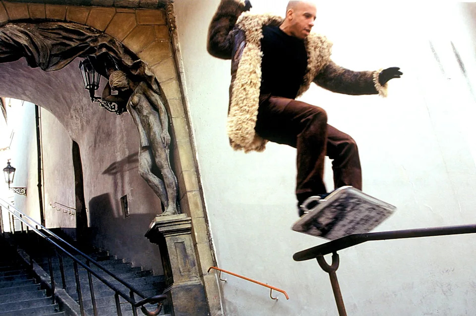 A person skateboarding down a stair railing, wearing a fluffy coat and dark pants, in an architectural setting with statues and street lamps
