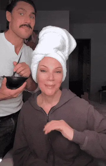 Person with wrapped hair and a makeup artist holding a brush and bowl, applying makeup on her face