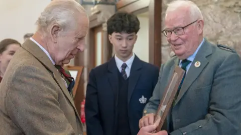 PA Media King Charles III, who has white hair and is wearing a brown tweed jacket, is presented with the framed score of a fiddle tune by Gordon Riddler, who also has white hair and is wearing a blue/green tweed jacket and a blue shirt with tartan tie. 