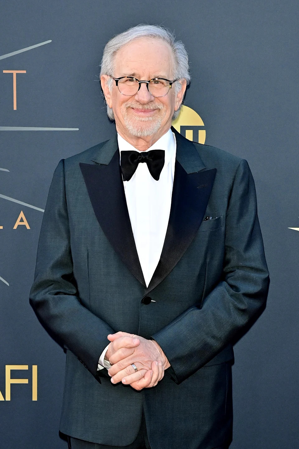 A person in a formal suit with a bowtie stands smiling at a celebrity event backdrop