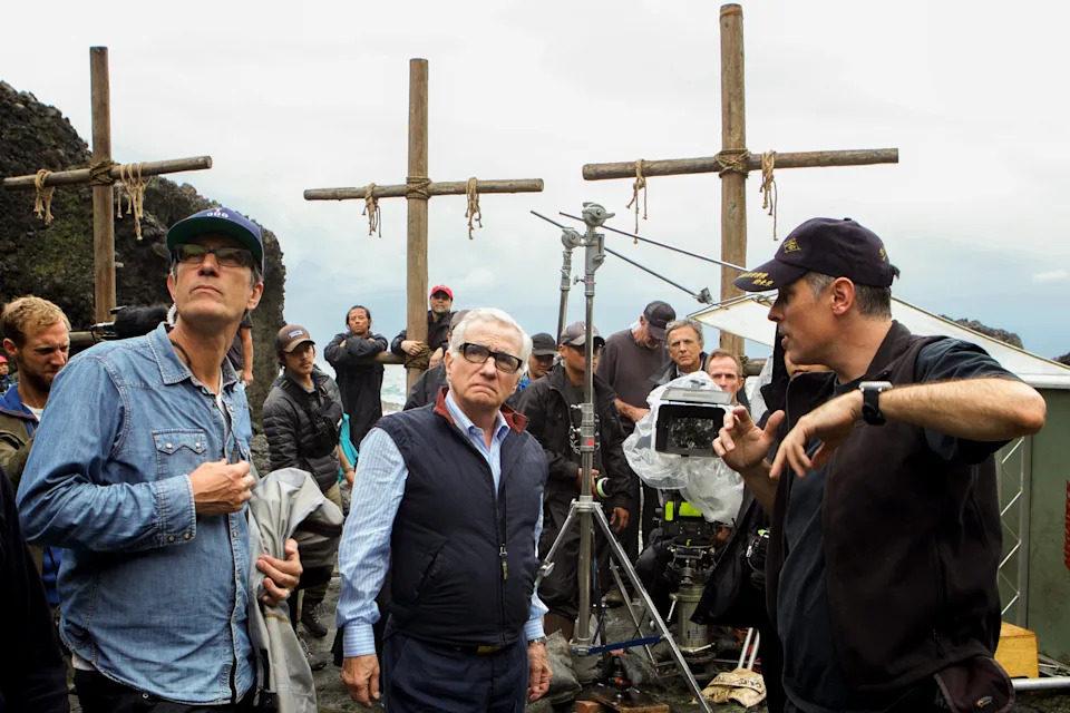 People on a film set with crosses in the background, wearing casual and work attire. One person holds a camera, others engage in discussion
