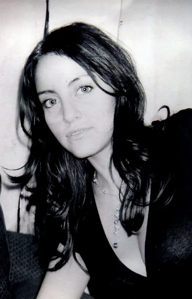 Person with long hair, wearing a necklace and V-neck top, looks towards the camera