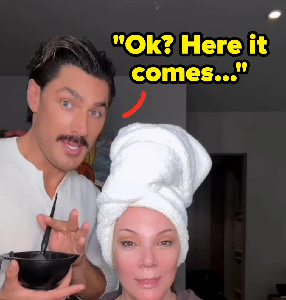 A person with a towel on their head and a man with a mustache smiling, holding a bowl with a brush, possibly in a salon or makeup setting