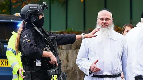 AFP via Getty Images Armed police officer talk to Rabbi Walker in front of a wall with green fencing above
