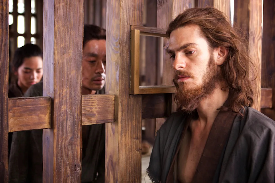 Two individuals peer through wooden bars at a bearded man inside. The scene suggests tension or contemplation