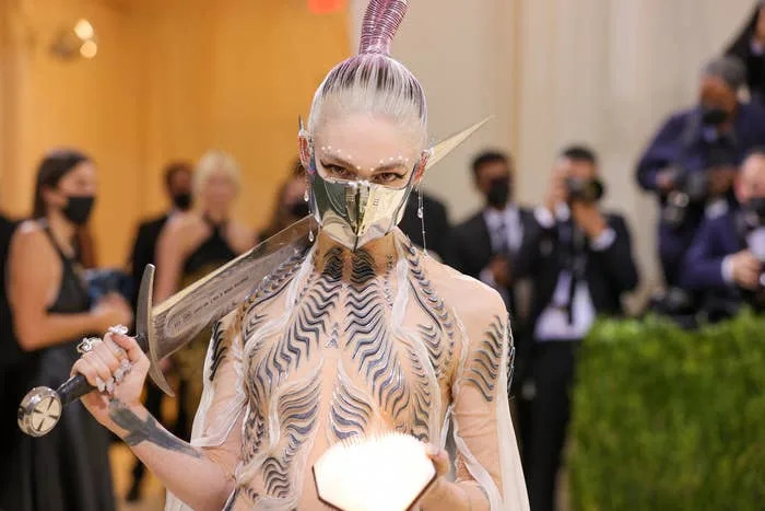 Person wearing an avant-garde outfit with metallic mask and carrying an ornate sword, posing for photographers at a high-profile event