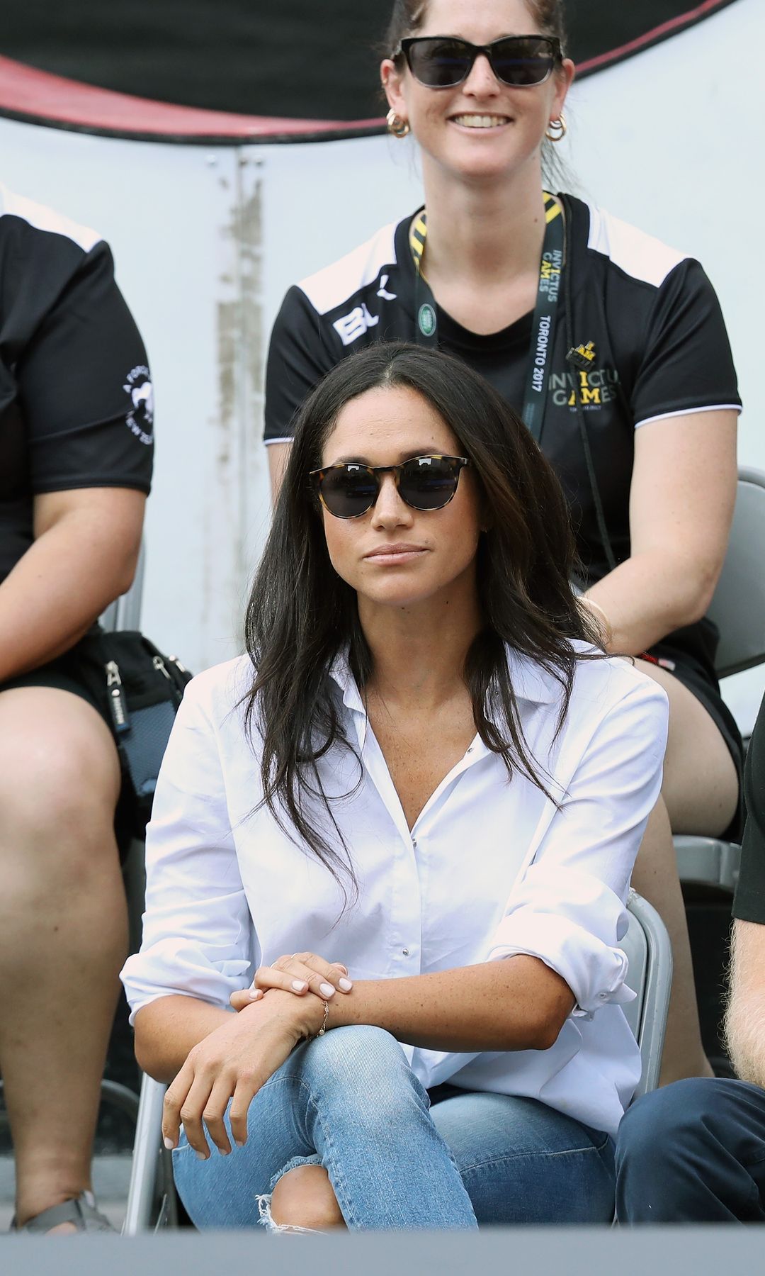 Prince Harry and Meghan Markle sit side by side watching the Invictus Games. Both wear jeans, Meghan teams hers with a white shirt and Harry with a black polo. They both wear sunglasses. 