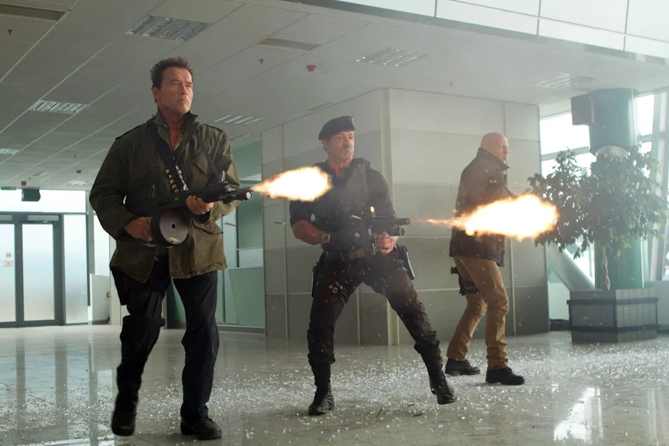 Three actors in military attire, armed and firing weapons, walk through a building with debris scattered on the floor