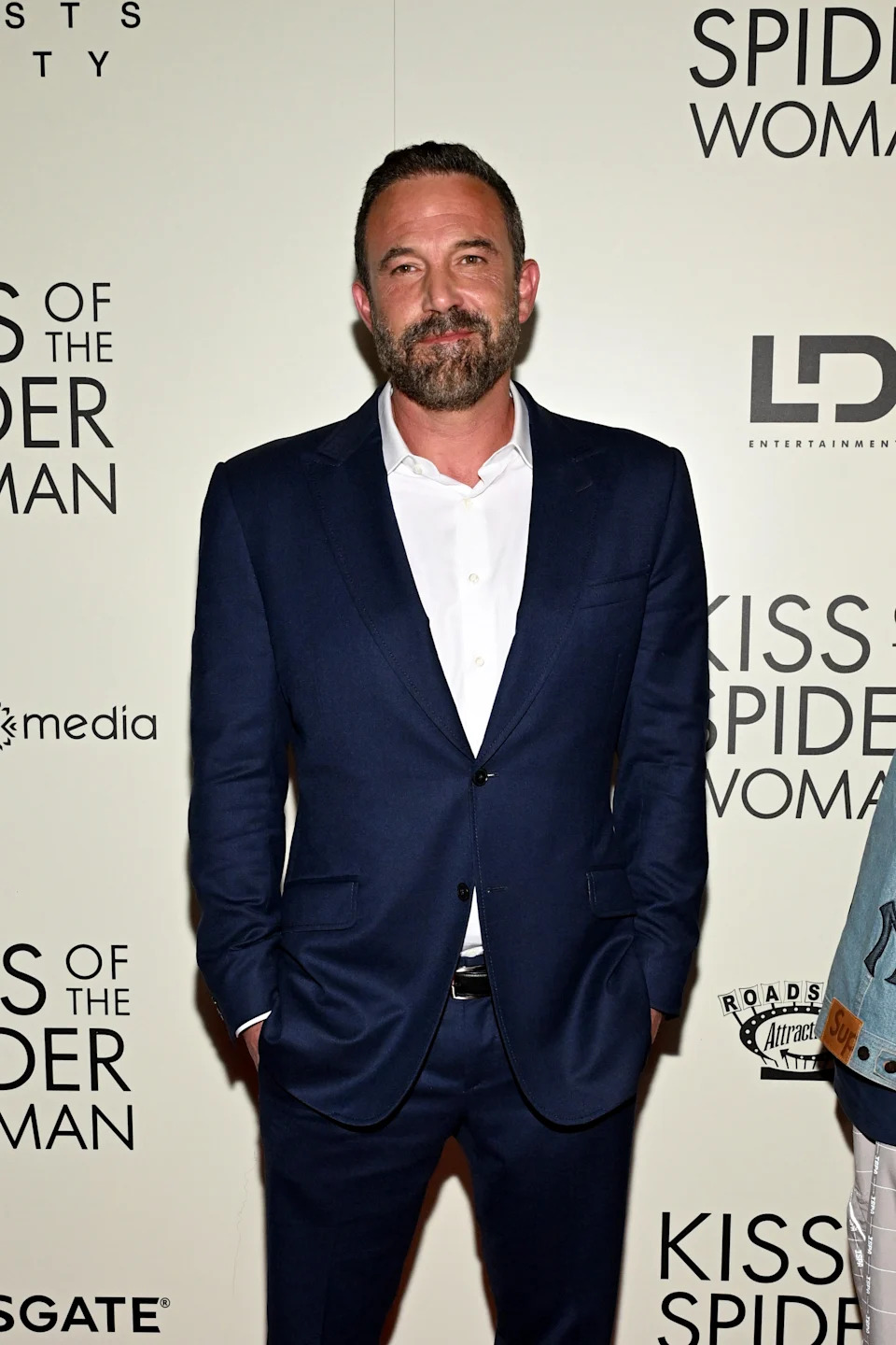 Man in a tailored suit at a premiere event, standing in front of a backdrop with text