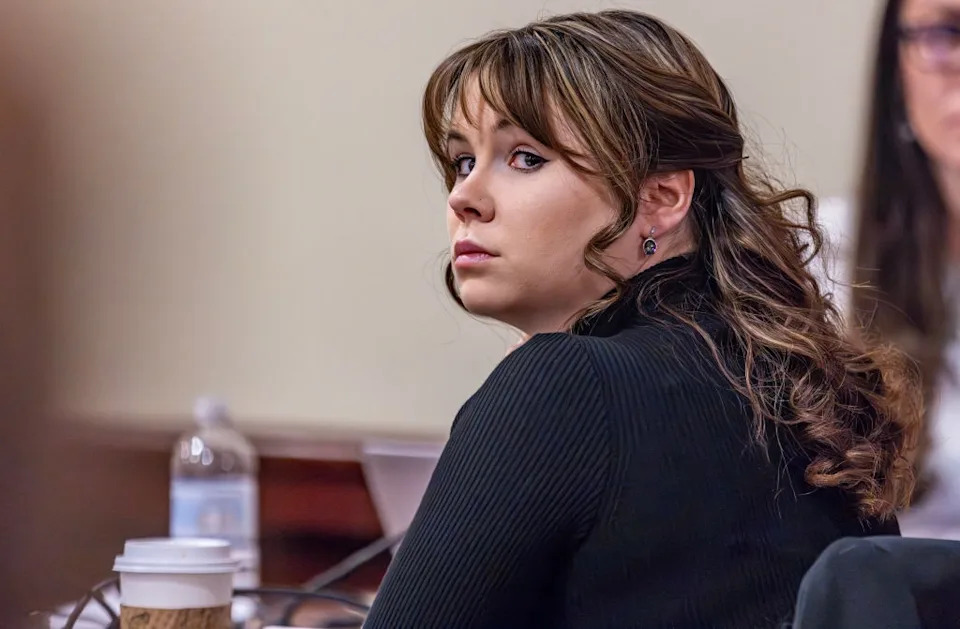 Woman with wavy hair in a dark turtleneck, seated at a table with a coffee cup and water bottle, looking back attentively