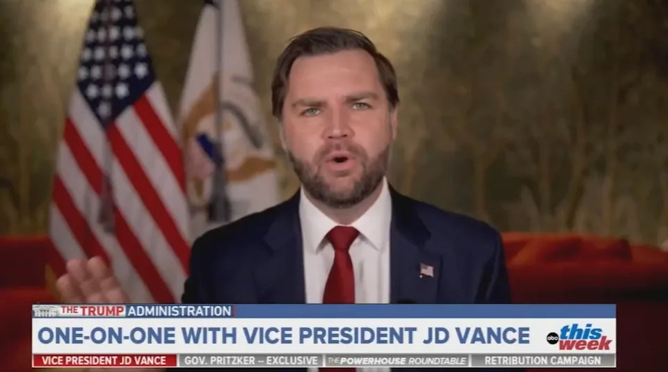 A man in a suit speaks during a TV interview with U.S. flags in the background. Text mentions Vice President JD Vance and "The Trump Administration."